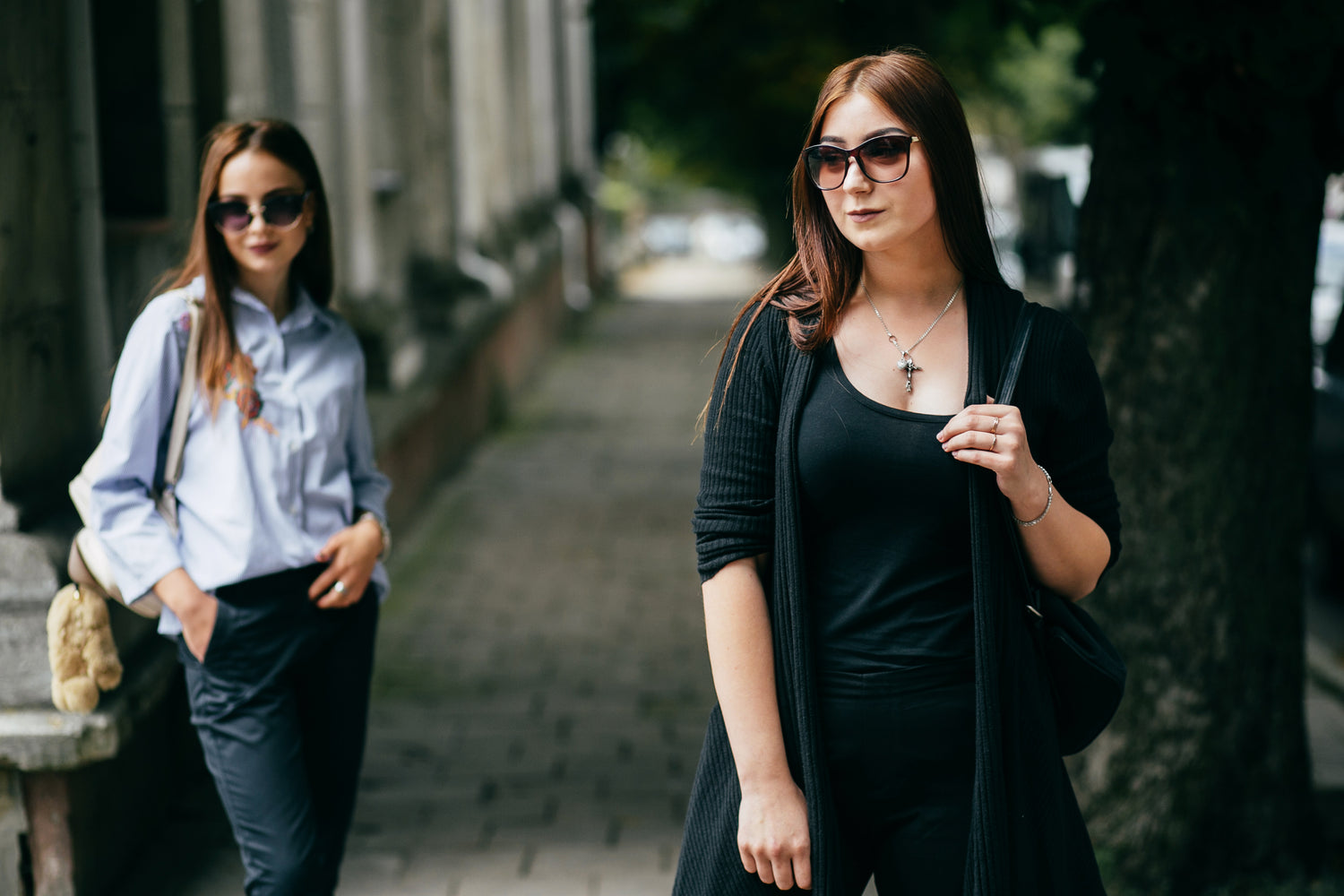 Two women walking on a street, one in a black outfit and sunglasses, the other in a light blue shirt and dark pants.